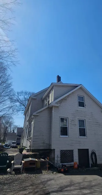 White two-story house with a steep roof and siding, viewed from the side under a clear blue sky.