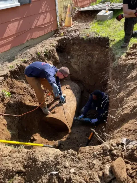 Two people work inside a deep excavated trench around a large, buried metal tank near a house.