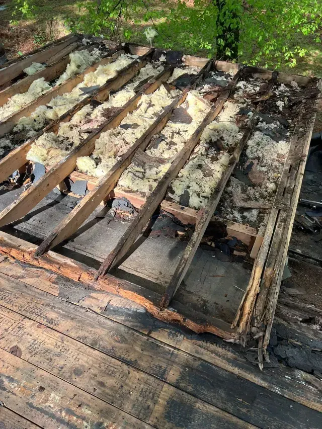 Damaged wooden deck with warped beams and missing boards, surrounded by debris and greenery