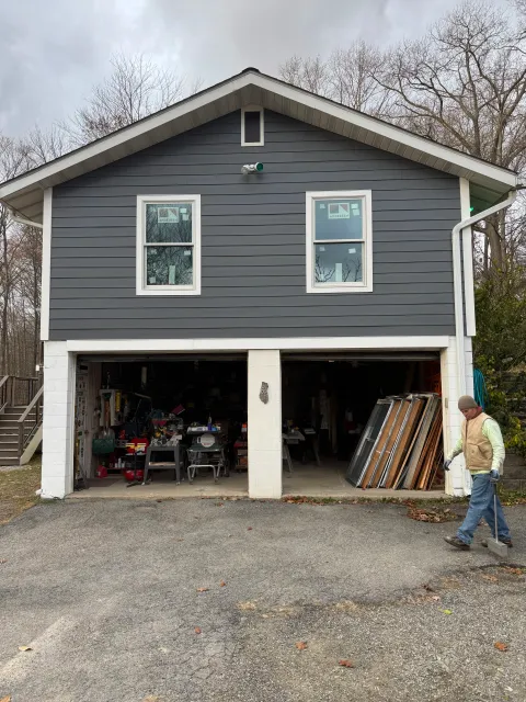 Two-car garage with the door open filled with tools and wood, a man in a vest and beanie walking outside. Bare trees and overcast sky in the background.