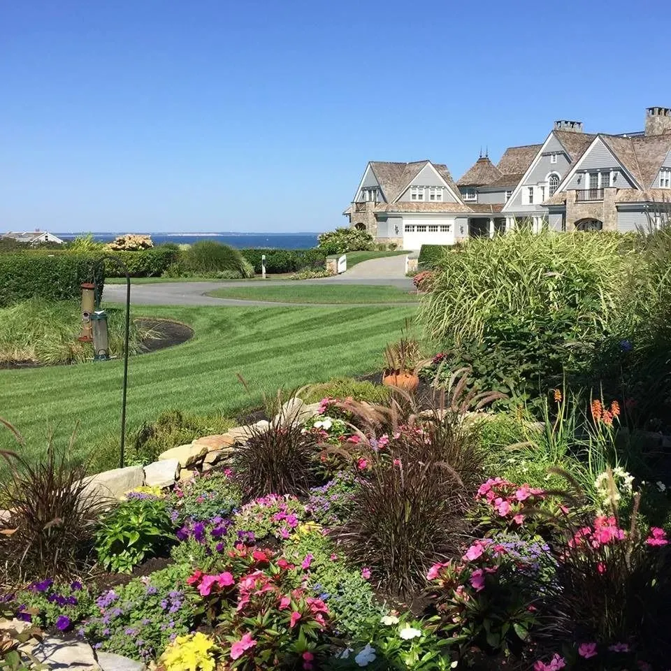 Lush garden in foreground with colorful flowers, green lawn with houses and ocean in background on a sunny day.