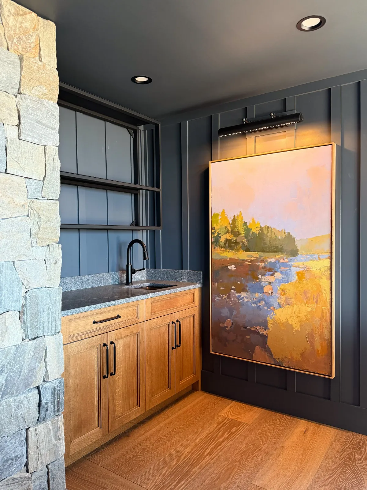 Hardwood floor installation in a Plymouth room featuring a stone accent wall, a blue painted wall with illuminated landscape artwork, a wooden cabinet with a gray countertop and black faucet, and natural wood flooring.