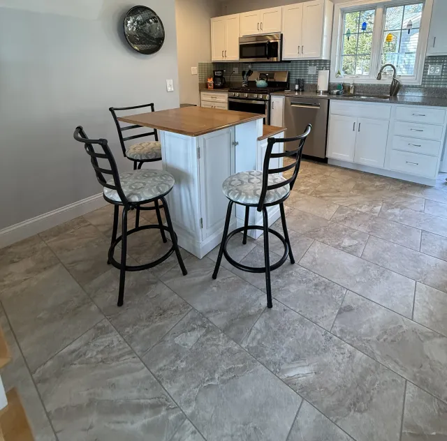 A modern kitchen featuring a wooden island with black metal bar stools, gray tiled flooring, and white cabinets. A stainless steel fridge and stove are visible in the background, along with a sink and a window letting in natural light.