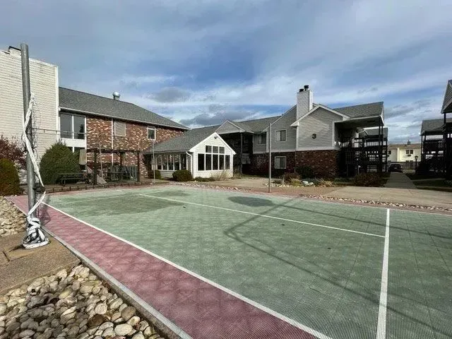 A green and maroon sports court with a net, next to buildings under a cloudy sky.