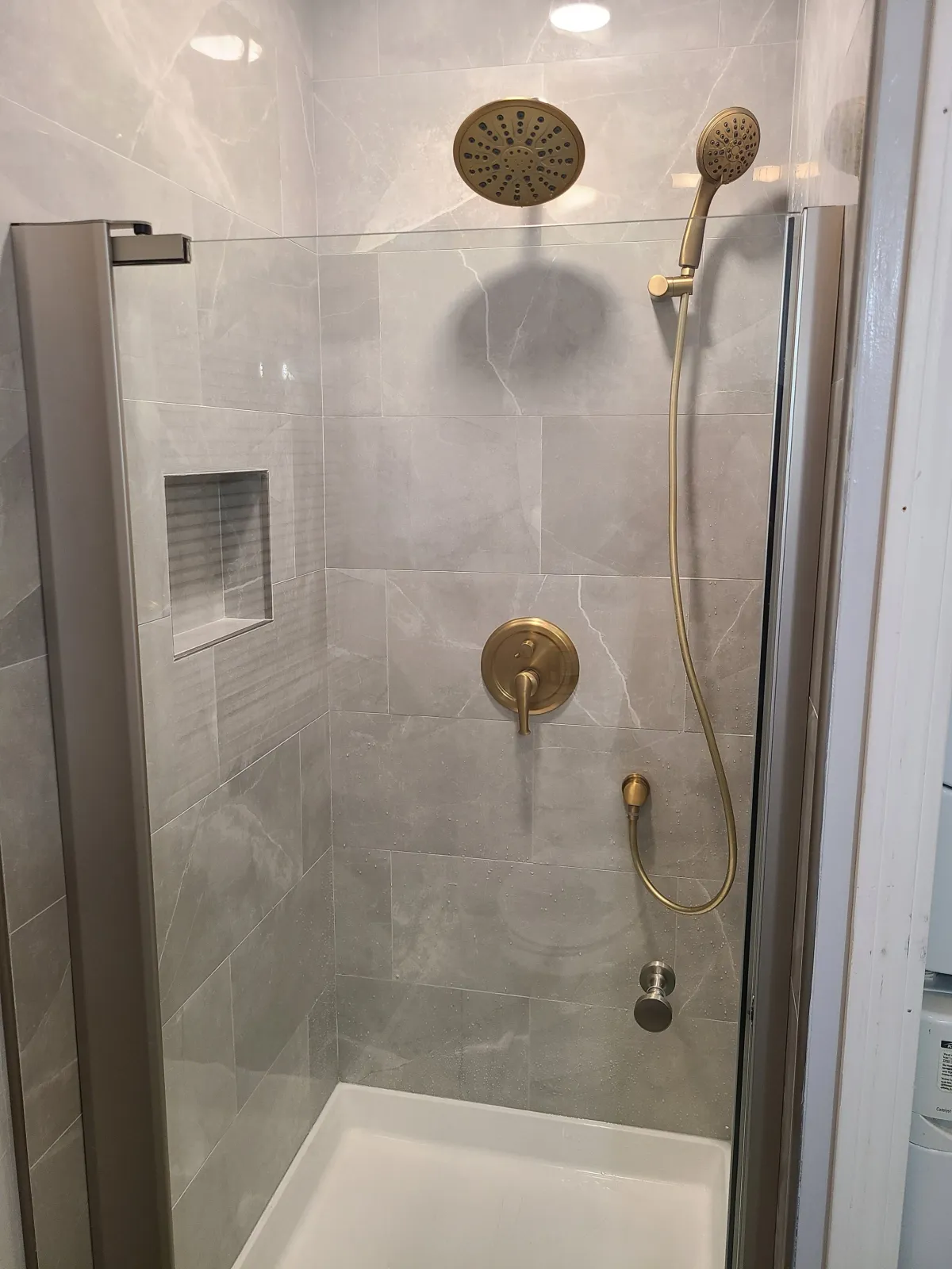 A modern shower with gray tiled walls, a glass door, a recessed soap niche, a brass showerhead, a hand-held shower wand, and a brass control panel.