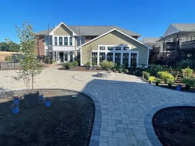 Backyard with paved patio, garden beds, and two-story house with light-green siding and large windows on a sunny day.