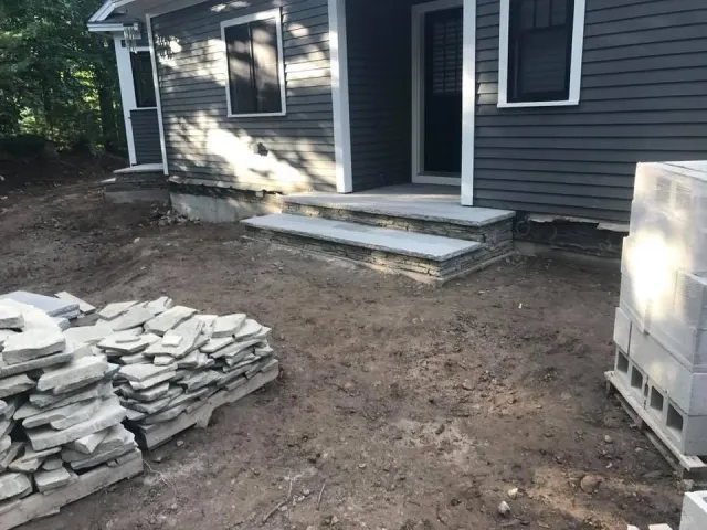 Construction site with stone steps leading up to a house entrance, and building materials stacked nearby.