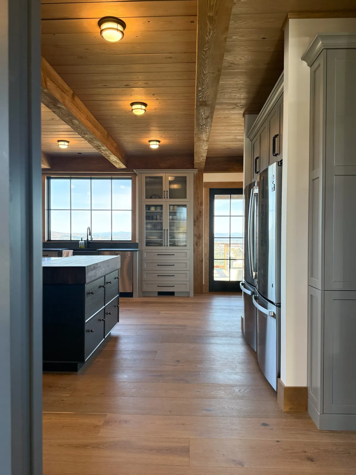 Hardwood floor installation in a Norwell modern kitchen with a wooden ceiling and floor, large window views, stainless steel refrigerator, white cabinetry with black handles, and a kitchen island with storage drawers.