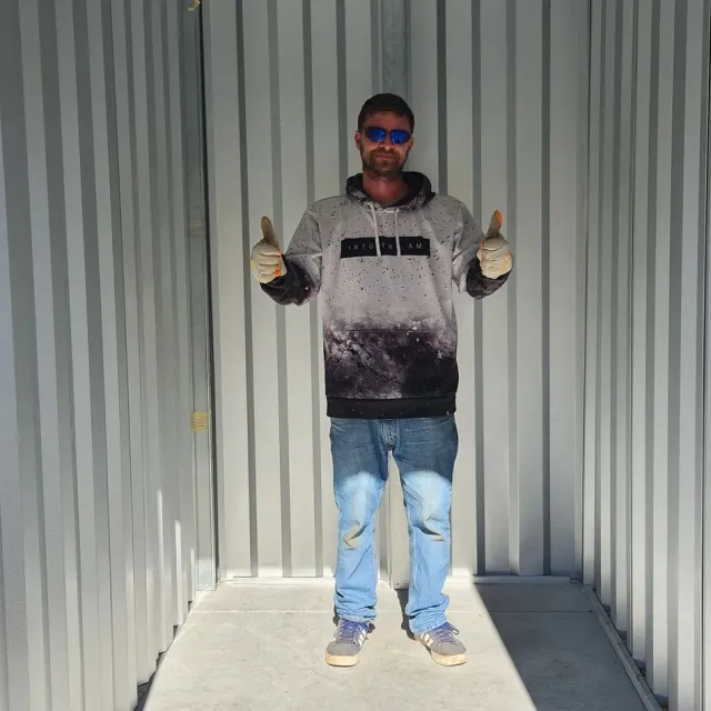 Person standing in a corrugated metal container, wearing sunglasses, thumbs up, in sunlight