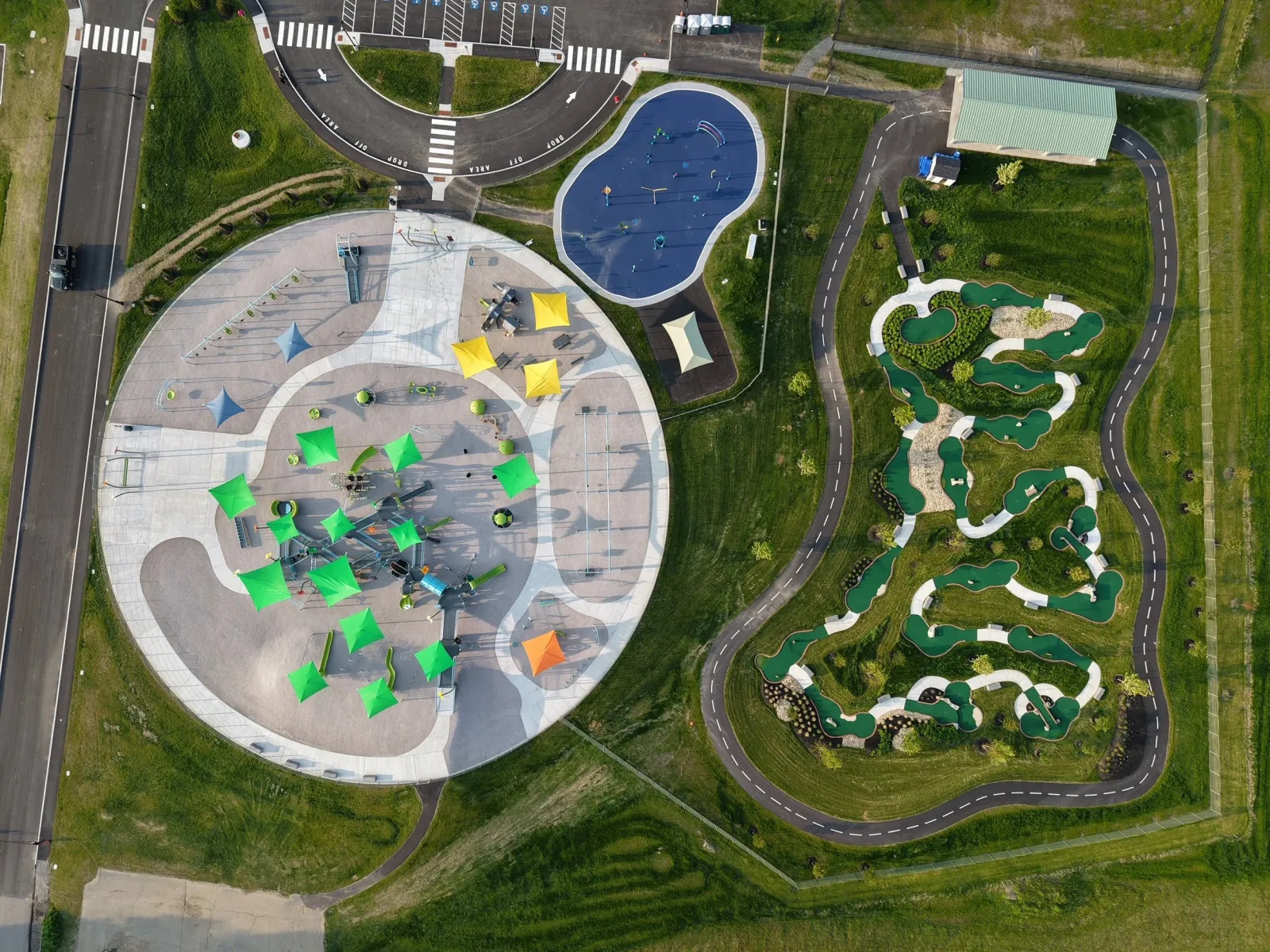 An aerial view of a park showing a playground with colorful tents, a blue splash pad, and a mini golf course with winding pathways surrounded by green grass.