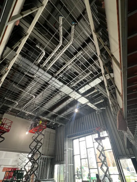 Construction site interior with high ceiling, visible ductwork, scaffolding lifts, and workers. Large windows provide natural light. Metal framing and unfinished walls are seen.