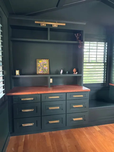 Dark green built-in cabinet with gold hardware, a wooden countertop, and a window with louvered blinds, with decorative items including a small clock, painting, penguin figurine, tree-shaped ornament, and shell, on the shelves.