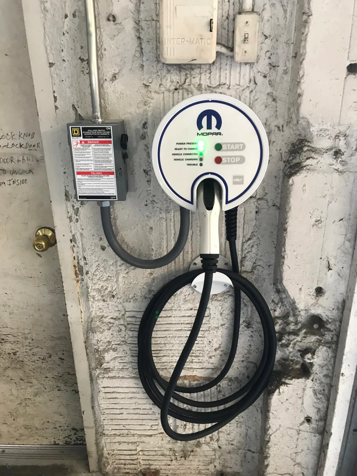 An electric vehicle charging station mounted on a concrete wall with a power outlet and electrical box nearby. The station has a circular display with indicators labeled 'Power Present', 'Ready to Charge', 'Vehicle Connected', 'Vehicle Charging', and