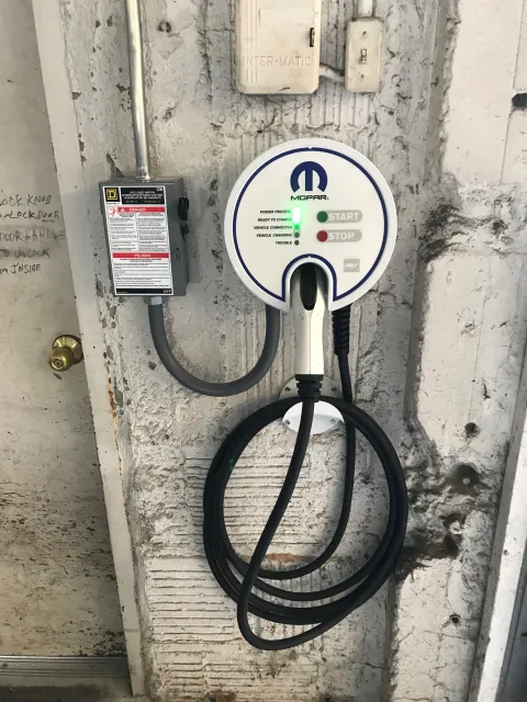 An electric vehicle charging station mounted on a concrete wall with a power outlet and electrical box nearby. The station has a circular display with indicators labeled 'Power Present', 'Ready to Charge', 'Vehicle Connected', 'Vehicle Charging', and