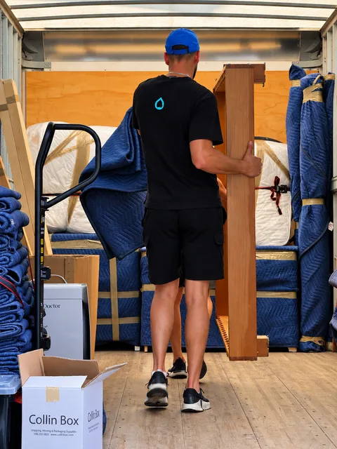 Mover in black tee leaning on blue padded furniture wrapped in plastic and tape inside a home.