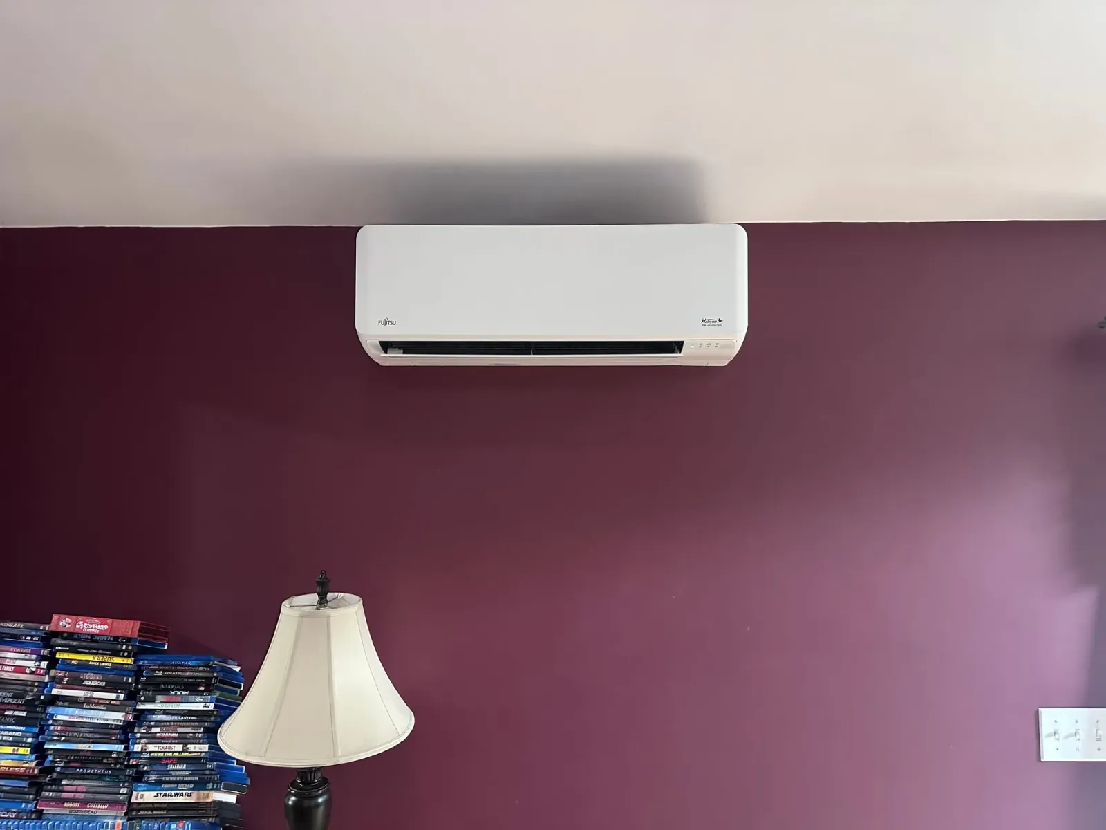 White wall-mounted air conditioner on a purple wall above a lamp and stacked items