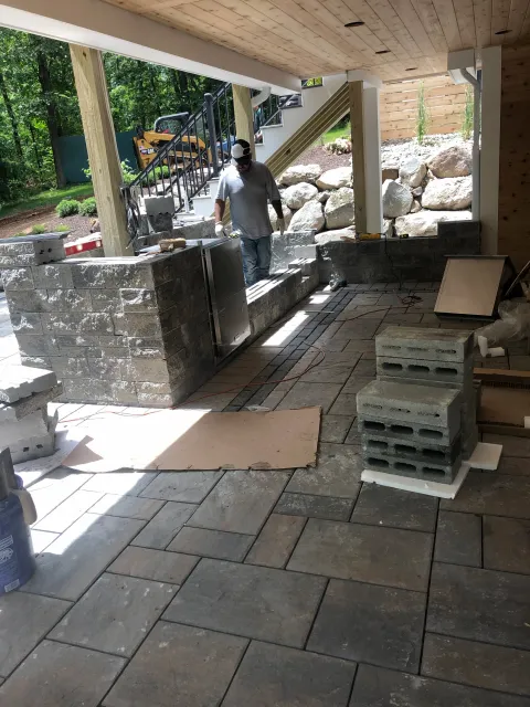 Construction worker installing a stone or brick countertop on an outdoor bar or kitchen area with stone flooring, under a covered patio with a staircase and landscaped yard in the background.