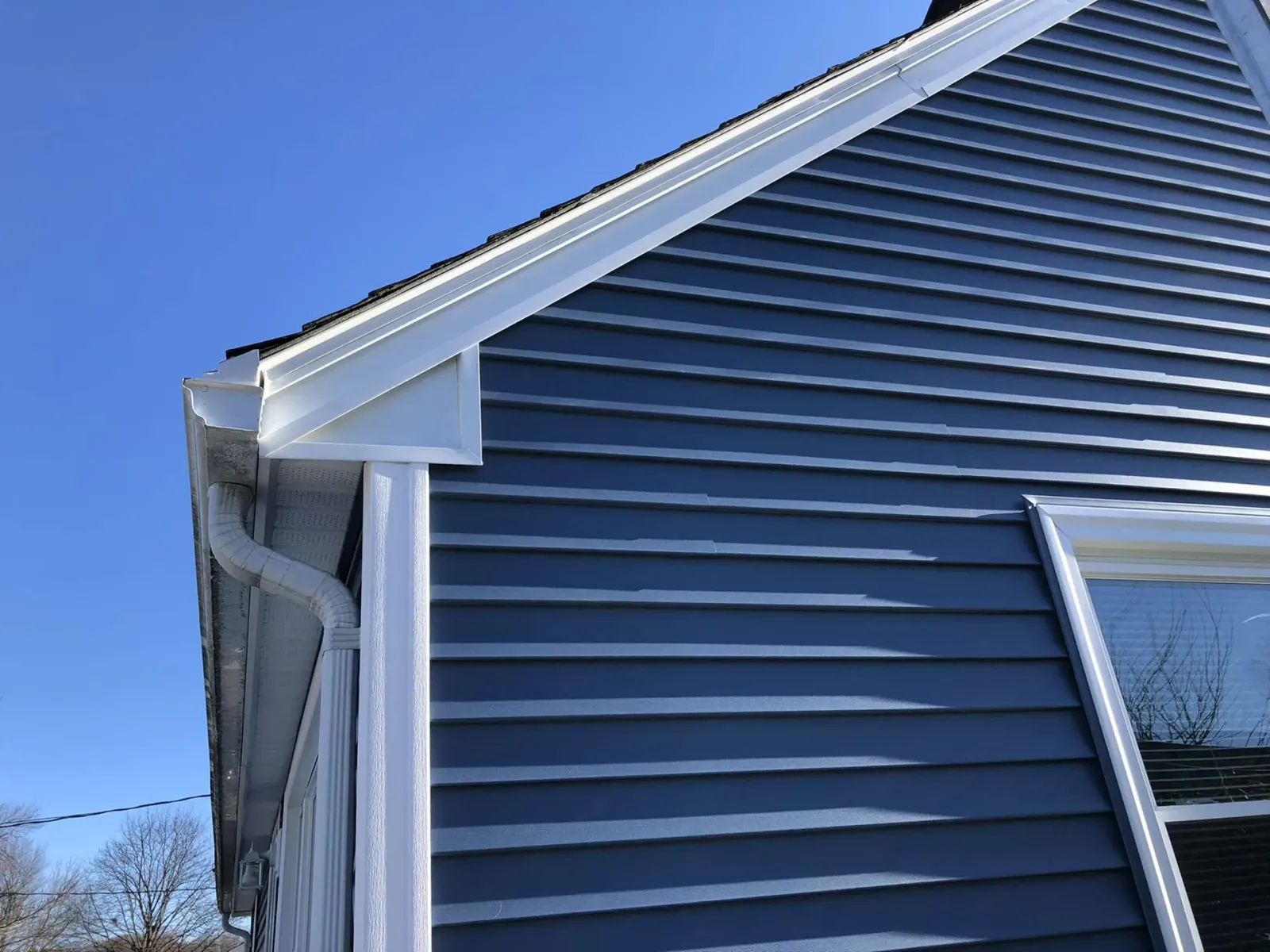 The corner of a blue-sided house with white trim, a gabled roofline, and an attached white gutter system against a blue sky.