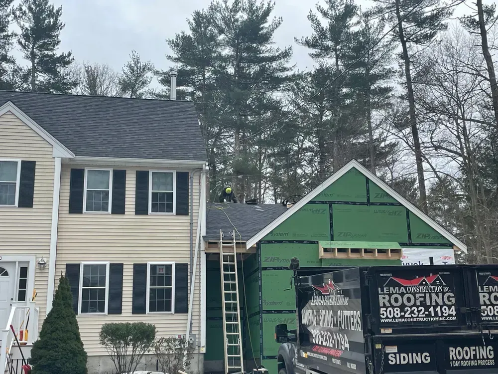 A house undergoing renovations with a worker on the roof, a ladder, and a parked roofing company truck in the driveway.