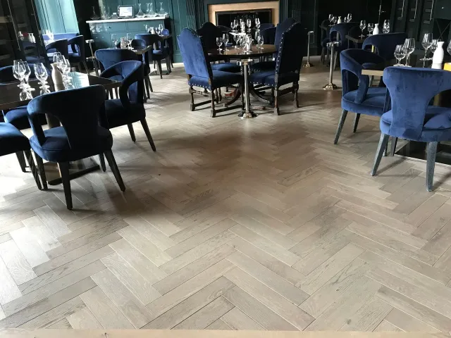 Restaurant interior with light wood parquet floor, blue velvet chairs, and round tables set with glasses.
