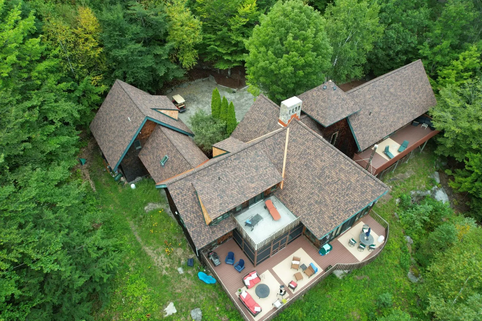 Aerial view of a rustic lodge with brown roofs surrounded by dense green forest, with a patio and tables outside