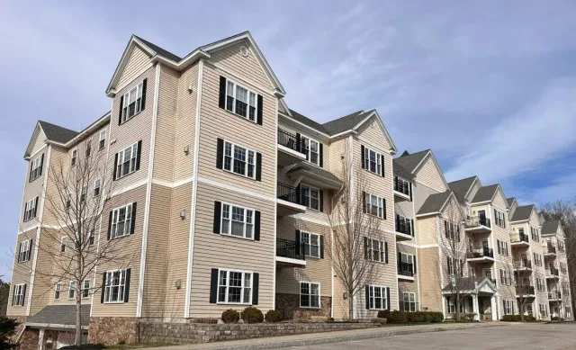 Multi-story apartment building with beige siding, black shutters, and balconies under a cloudy sky.