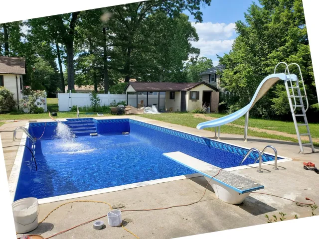 A backyard with an in-ground swimming pool and a waterslide. There is a small shed or pool house with a brown roof, and a white fence surrounding the yard. Tall trees with green leaves are in the background, and the sky is partly cloudy.