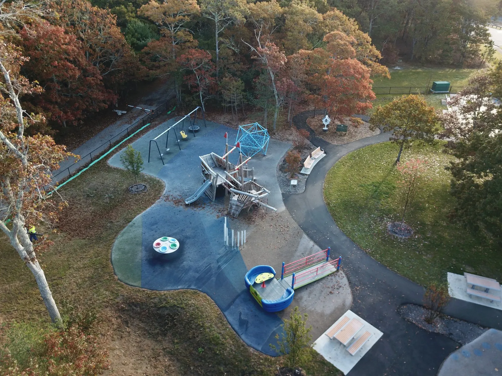 Aerial view of a playground with swings, slide, and climbing structure, surrounded by trees with fall foliage, walking paths, benches, and a grassy area.