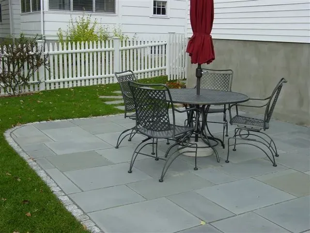 Patio with black metal table and chairs on gray pavers beside a white fence and house.