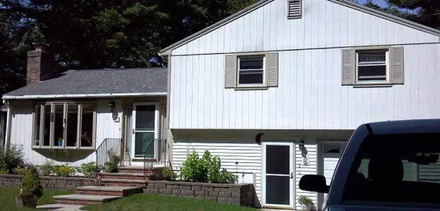 A white, two-story house with vertical siding, a bay window, and a stone retaining wall in the front yard.