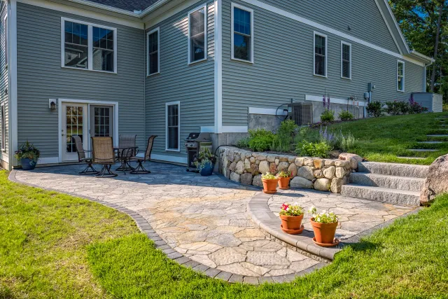 Backyard patio with stone paving, outdoor dining set, grill, stone retaining wall, and potted plants near a two-story house.