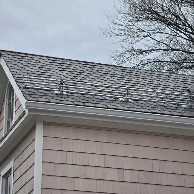 A metal snow guard rail installed on the dark gray shingled roof of a house with tan exterior siding.