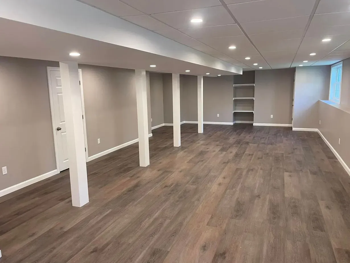 Empty finished basement room with beige walls, white columns, and wood-look flooring