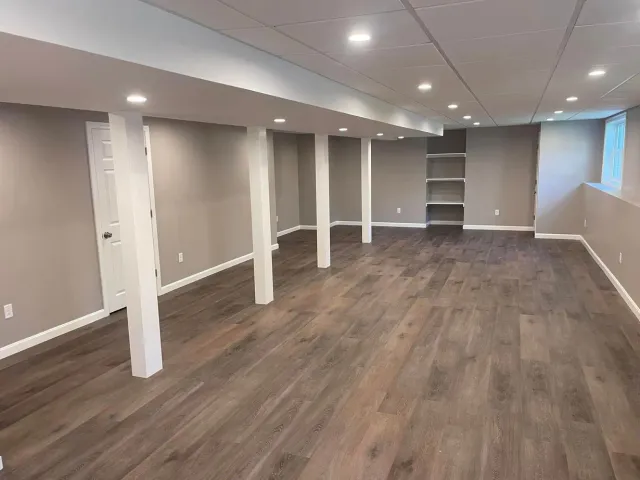 Empty finished basement room with beige walls, white columns, and wood-look flooring