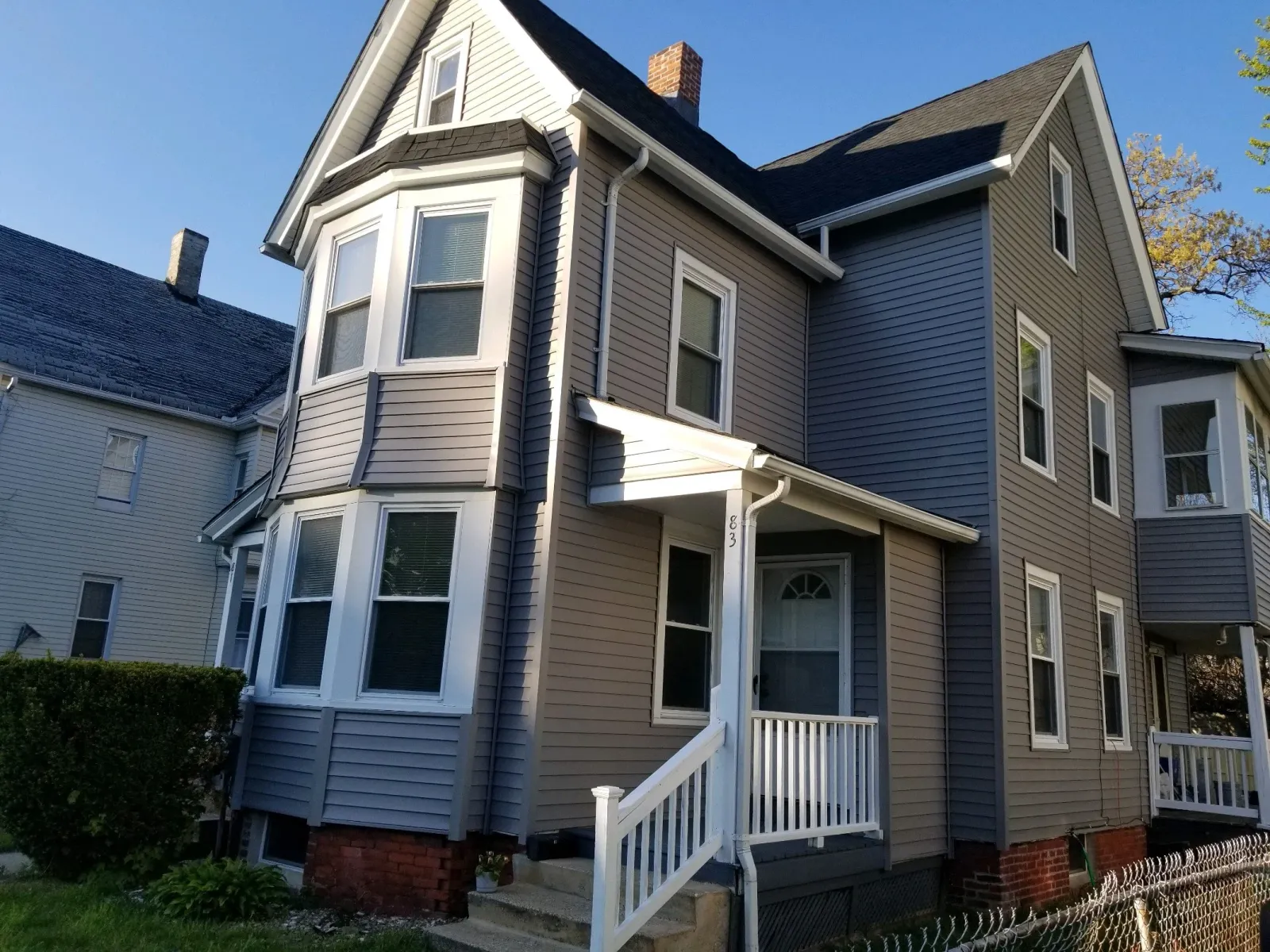 A three-story house with beige siding, experienced sunlight, a white front porch with stairs, and multiple windows, including a prominent bay window on the front.