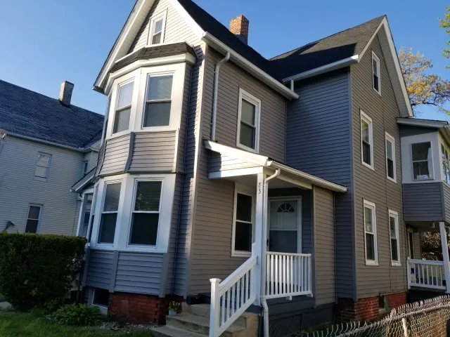 A three-story house with beige siding, experienced sunlight, a white front porch with stairs, and multiple windows, including a prominent bay window on the front.