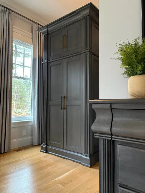 A corner of a room with a window covered by beige curtains, a black wardrobe with gold handles, a potted plant on the dresser, and hardwood flooring.