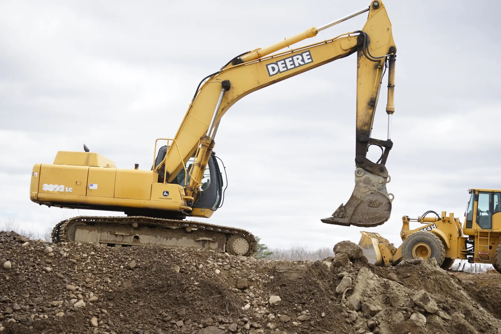 A large yellow John Deere excavator digging into a dirt mound on a construction site under a cloudy sky.