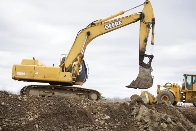 A large yellow John Deere excavator digging into a dirt mound on a construction site under a cloudy sky.