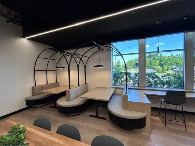 Modern dining area with curved booth seating, black metal arch structures, pendant lighting, large window showing green trees and blue sky, and a black chair near the window.
