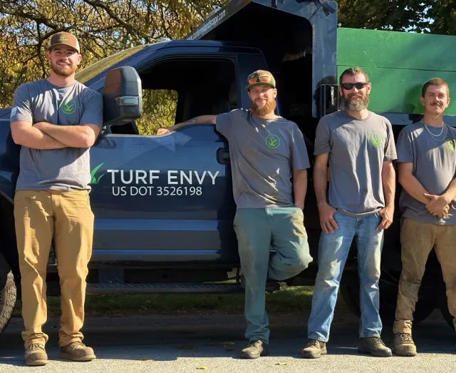 Five men standing in front of a large truck with the logo 'Turf Envy' on the side, outdoors during daytime. They are all wearing gray t-shirts with the logo, and appear to be workers or contractors.
