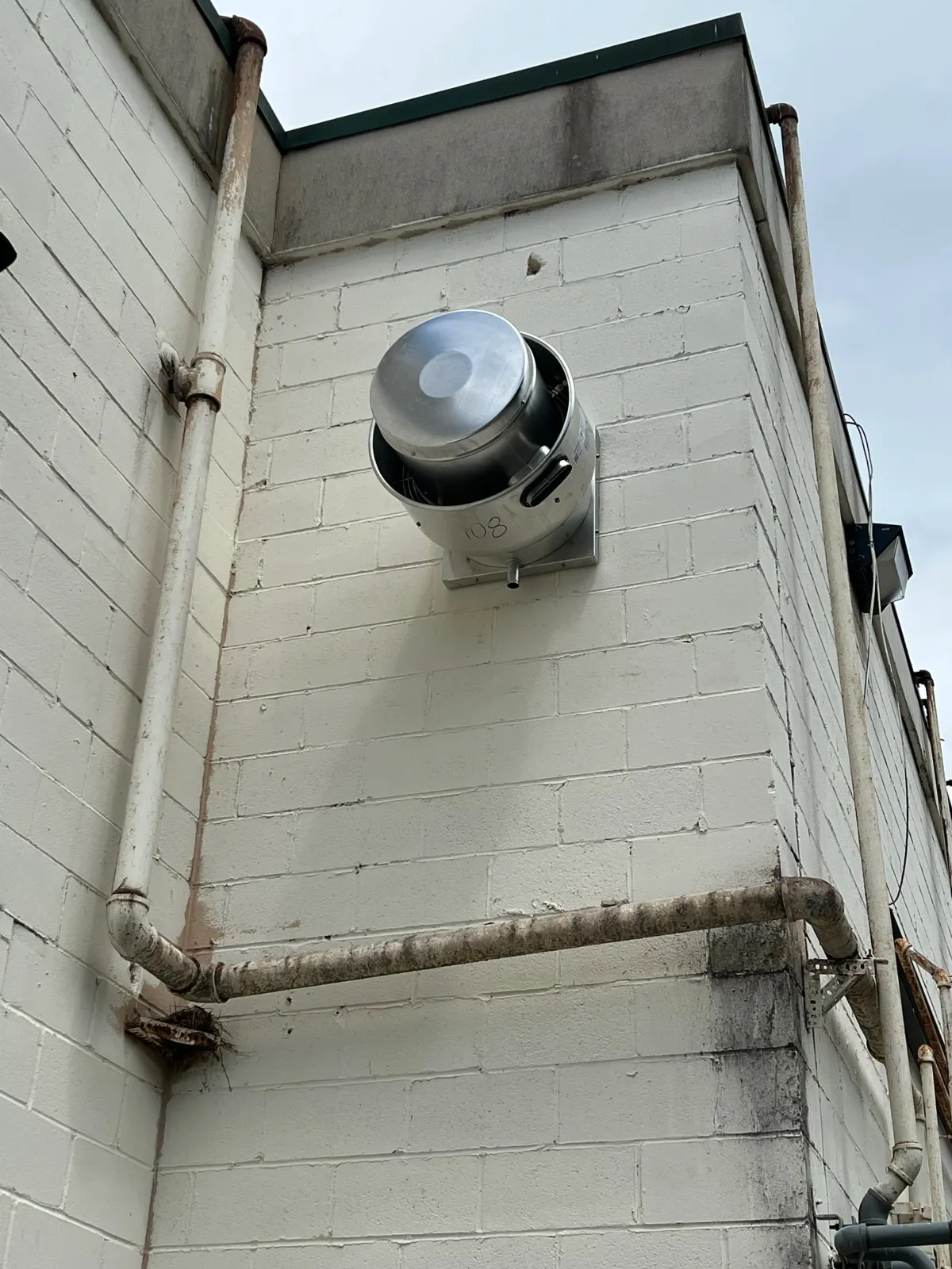 An exterior corner of a building with cinder block walls and a satellite dish mounted on the wall. There are metal pipes running along the wall, some showing rust and wear.