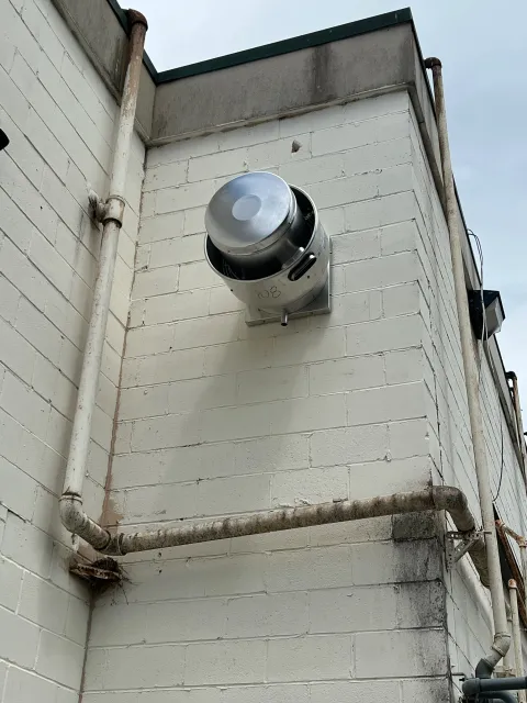 An exterior corner of a building with cinder block walls and a satellite dish mounted on the wall. There are metal pipes running along the wall, some showing rust and wear.