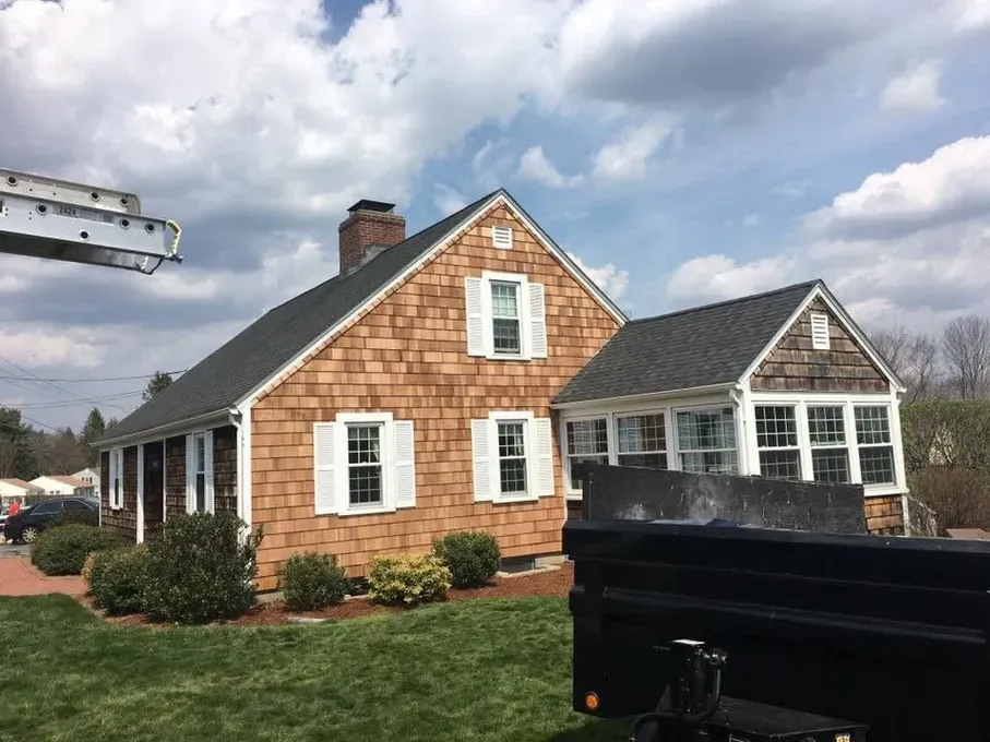 Brick house with white trim and dark roof under cloudy sky, with a black dumpster in front