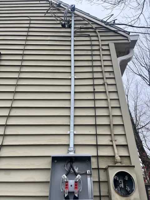 The exterior of a house with beige vinyl siding, showing electrical wiring and a metal conduit running vertically. There is an electrical box with wires inside, and a circular electric meter on the side of the house. The house has a roofline with a gutter and some leafless tree branches are visible against the sky.