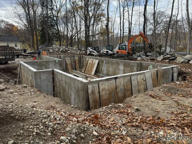 Foundation construction site; concrete walls are in place. An excavator and other equipment are visible.