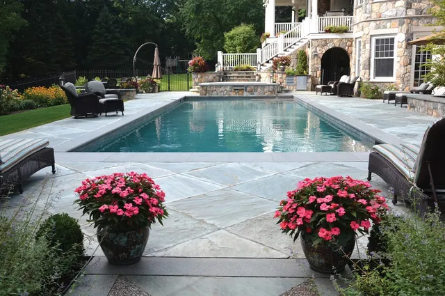 An outdoor swimming pool surrounded by stone tiles with patio furniture, lush landscaping, and a house with stone facade in the background.