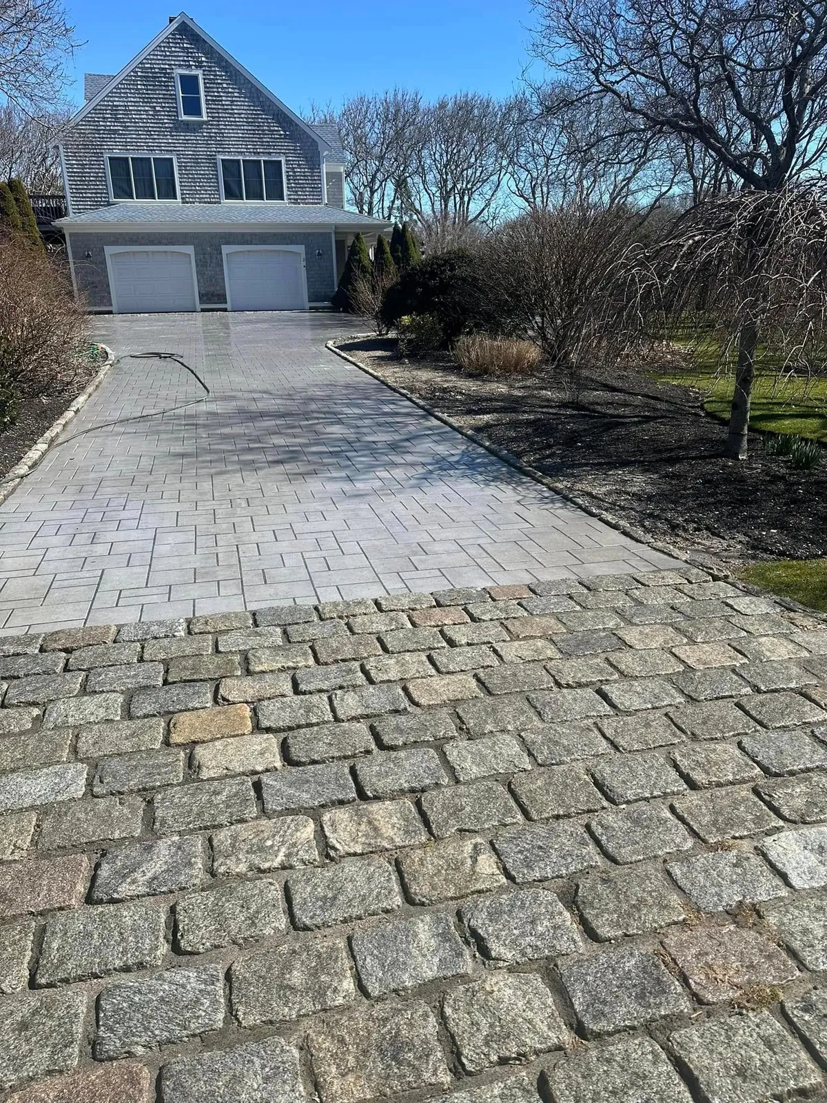 Driveway of a gray house, transitioning from cobblestone to paving stones. Sunny day.