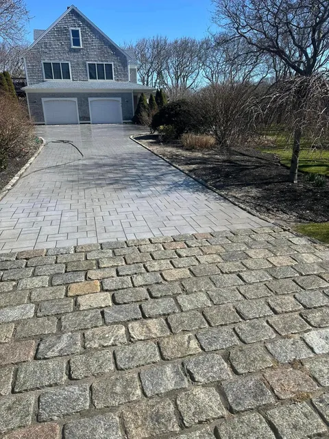 Driveway of a gray house, transitioning from cobblestone to paving stones. Sunny day.