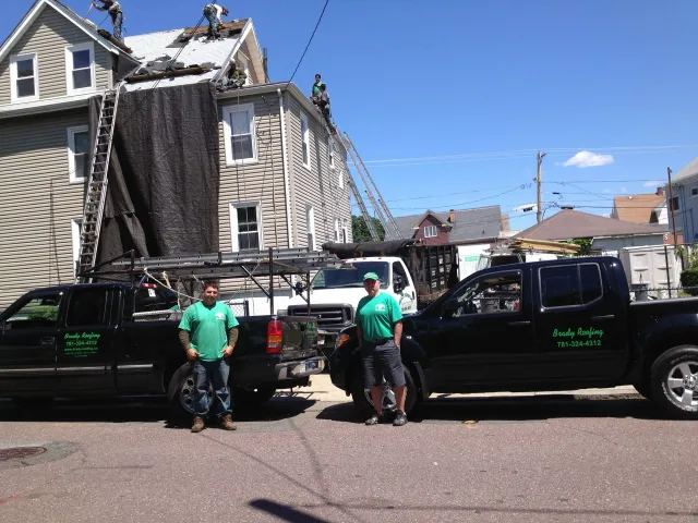Two workers beside black pickup trucks on a street, with a damaged house and debris in the background